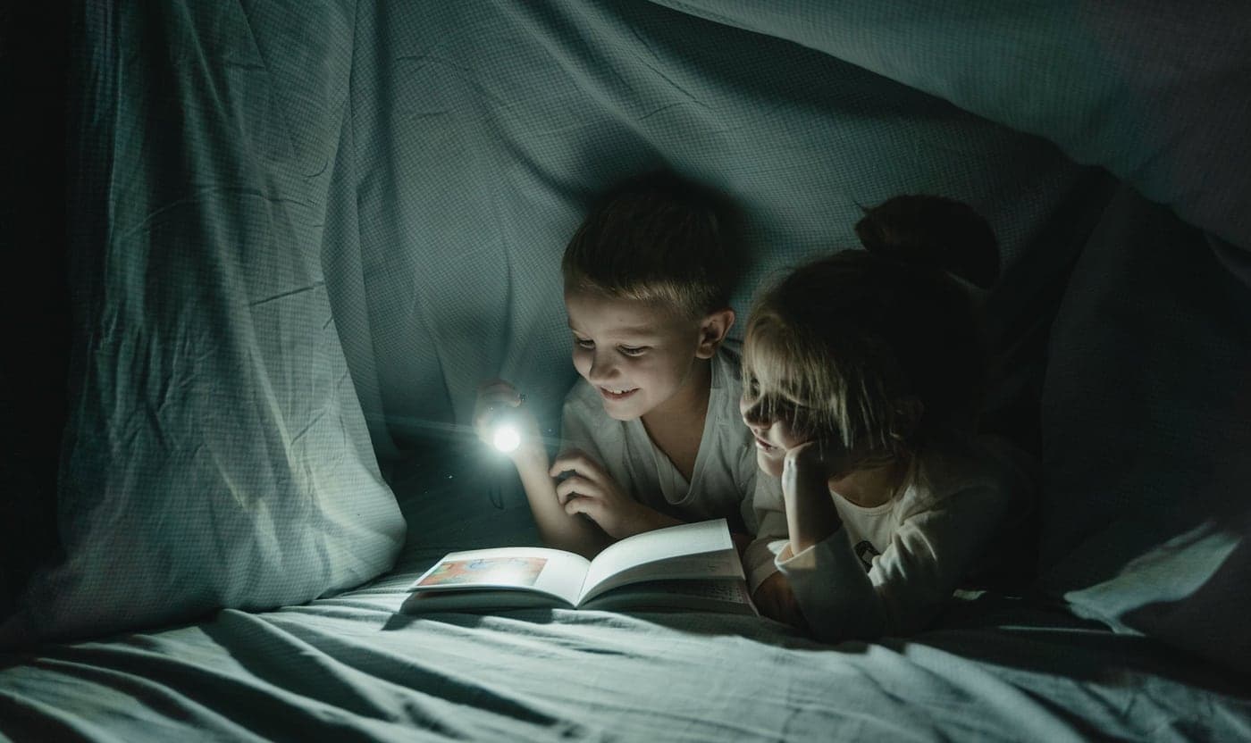 Two children lying in a blanket fort at night, sharing a picture book by torchlight